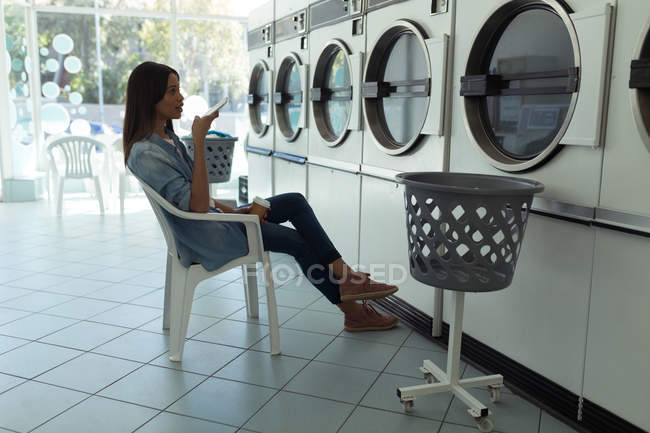 Jovem mulher falando ao telefone enquanto espera na lavanderia — Fotografia de Stock