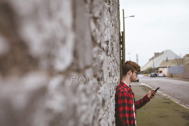 Jeune homme utilisant son téléphone portable contre un mur de pierre près de la rue — Photo de stock