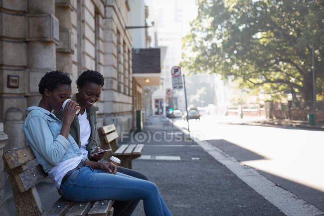 Fratelli gemelli che utilizzano il telefono cellulare mentre si rilassano sulla panchina in città — Foto stock