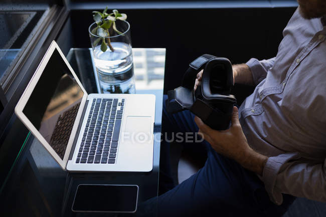 Businessman holding virtual reality headset while working on laptop at office — Stock Photo