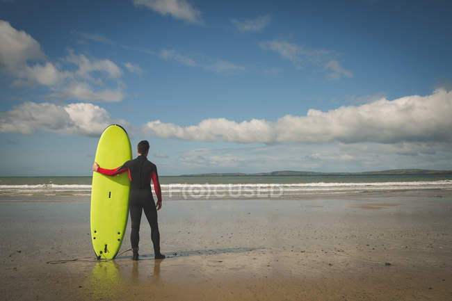Vista trasera del surfista con tabla de surf mirando al mar desde la playa - foto de stock