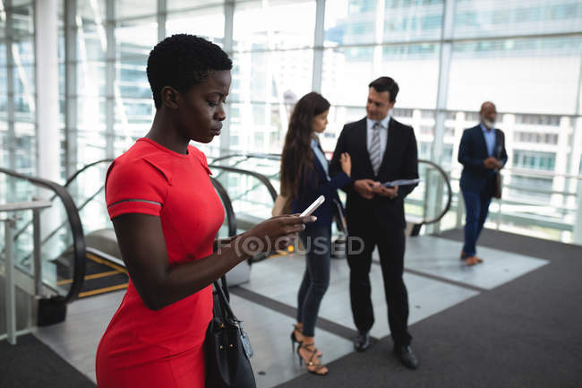 Бізнес-леді, використовуючи свій телефон, поки колеги обговорюють в офісі — стокове фото