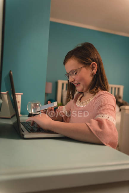 Girl looking at mobile phone while using laptop in bedroom at home — Stock Photo