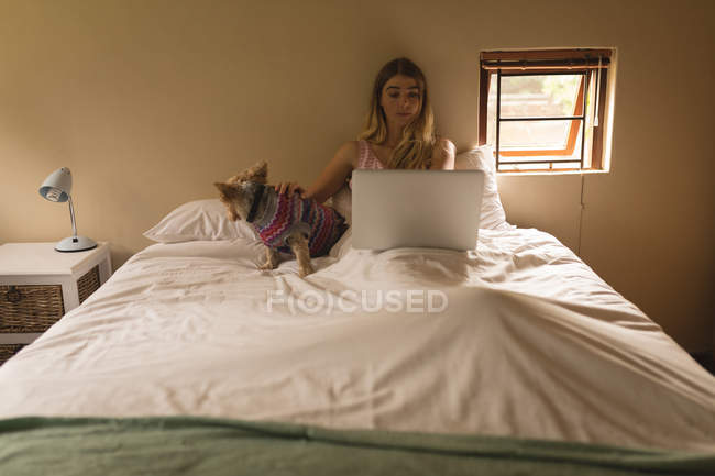 Дівчина з собакою використовує ноутбук у спальні вдома — стокове фото