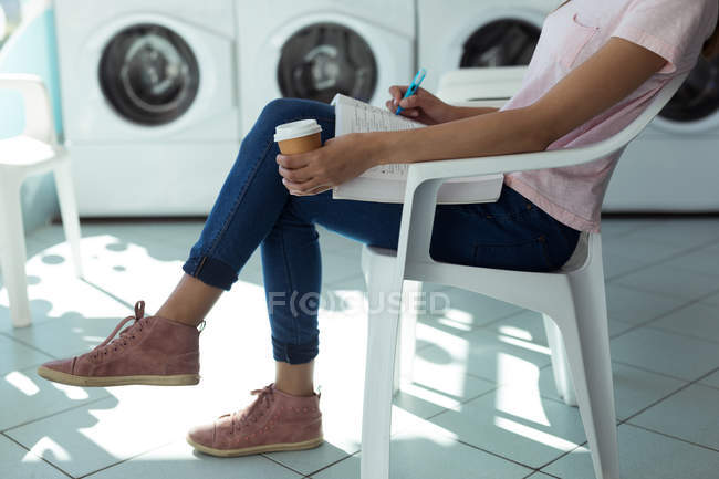 Low section woman with coffee cup writing in the book at laundromat — Stock Photo
