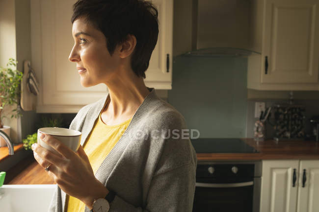 Femme regardant ailleurs tout en prenant un café dans la cuisine à la maison — Photo de stock