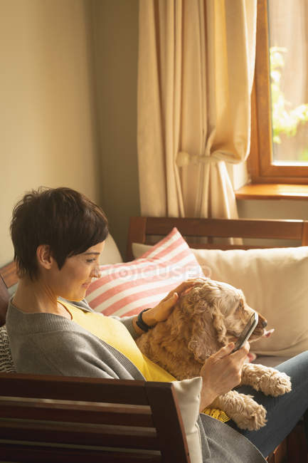 Mujer usando teléfono móvil con perro en el sofá en la sala de estar en casa - foto de stock