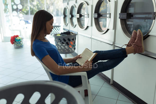 Jeune femme lisant un livre à la laverie automatique — Photo de stock