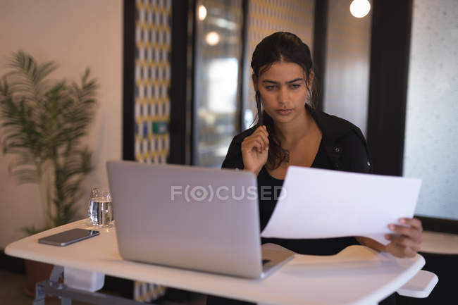 Geschäftsfrau liest Dokumente, während sie im Büro am Laptop arbeitet — Stockfoto