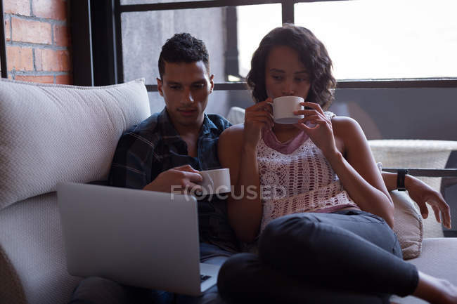 Pareja tomando té de limón mientras usa el portátil en la sala de estar en casa - foto de stock