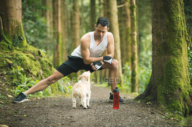 Здоровый человек делает упражнения на растяжку со своей собакой в лесу — стоковое фото