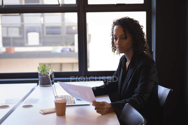 Mujer de negocios leyendo documentos mientras trabaja en el ordenador portátil en la oficina - foto de stock