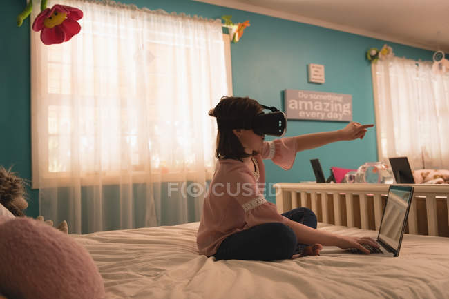 Fille en utilisant un casque de réalité virtuelle avec ordinateur portable dans la chambre à coucher à la maison — Photo de stock