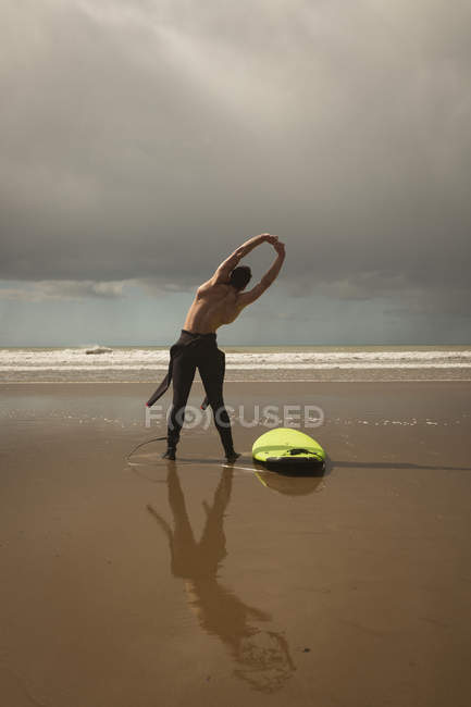 Rear view of surfer with surfboard performing stretching exercise at beach — Stock Photo