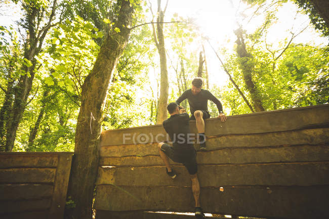Hombre en forma ayudando a su compañero de equipo a escalar una pared de madera en el campamento de entrenamiento - foto de stock