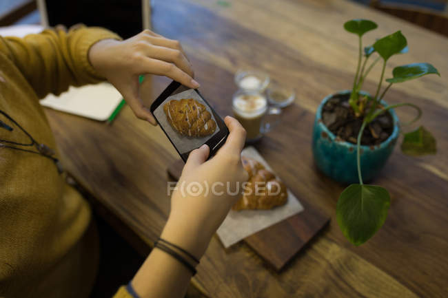 Молодая женщина фотографирует круассан, подаваемый на стол в кафе — стоковое фото