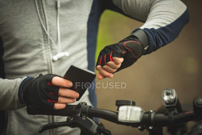 Sección media del ciclista comprobando el tiempo mientras usa el teléfono móvil en el bosque - foto de stock