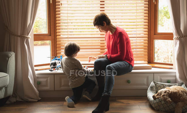 Madre jugando con su hijo en el alféizar de la ventana en la sala de estar en casa - foto de stock