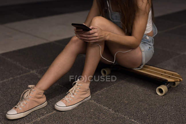 Жіночий ковзаняр сидить на скейті і використовує мобільний телефон на вулиці — стокове фото