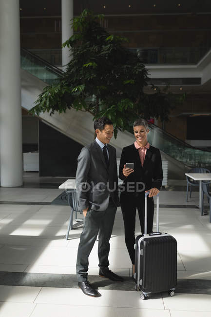 Улыбающиеся деловые люди с чемоданом смотрят на телефон в офисе — стоковое фото
