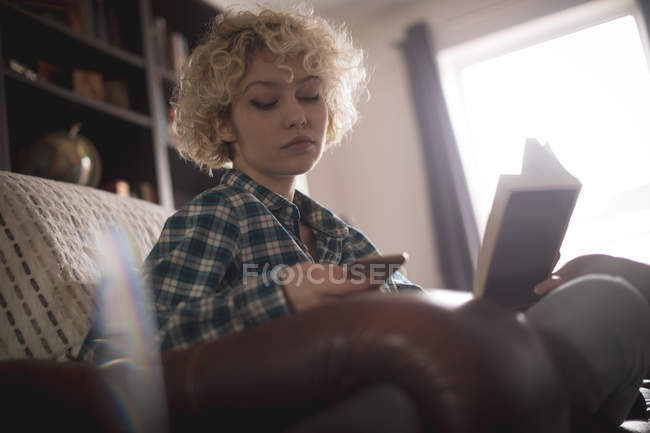 Жінка використовує мобільний телефон під час читання книги у вітальні вдома — стокове фото