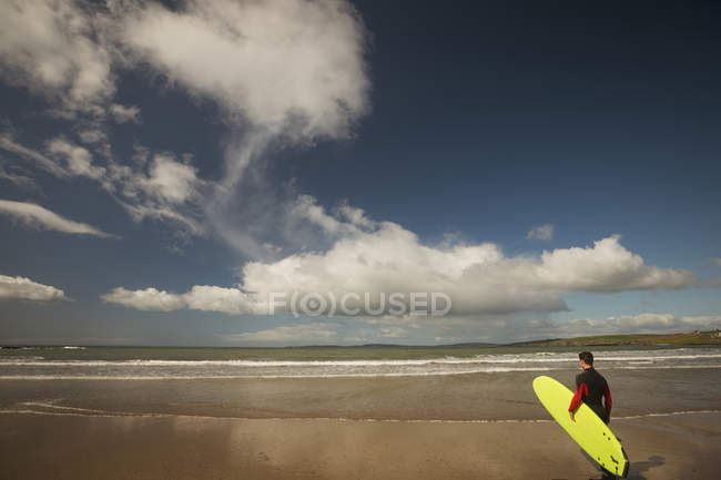 Surfista con tavola da surf che guarda il mare dalla spiaggia in una giornata di sole — Foto stock