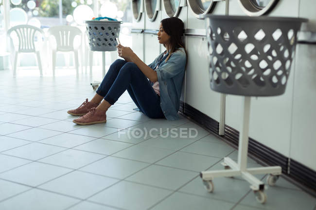 Mujer joven usando su teléfono mientras espera en la lavandería - foto de stock