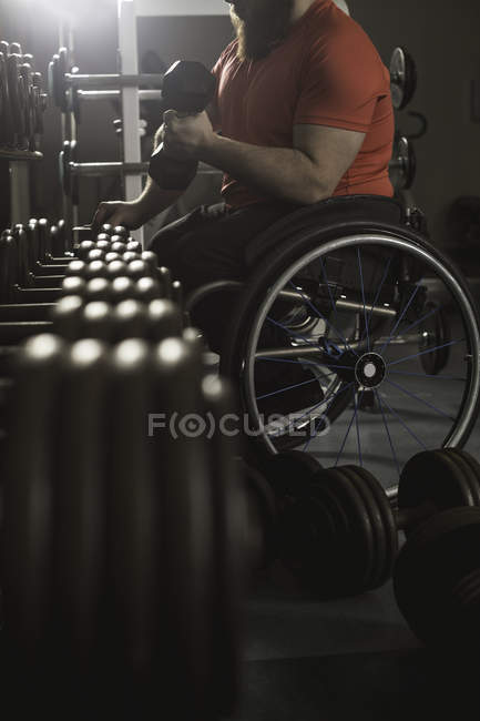 Handicapped man on wheelchair lifting dumbbell from rack in gym — Stock Photo