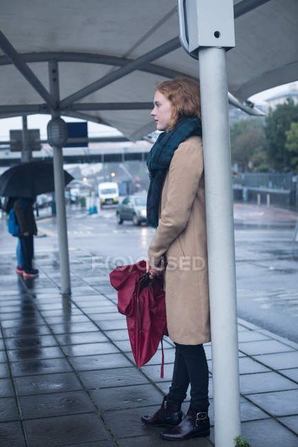 Молодая женщина держит зонтик стоя на автобусной остановке в дождливый день — стоковое фото