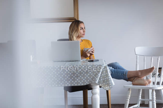 Задумчивая женщина с поднятыми ногами пьет кофе дома — стоковое фото