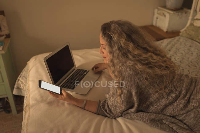 Зрелая женщина с помощью мобильного телефона и ноутбука в спальне дома — стоковое фото