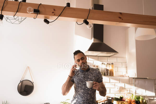 Мужчина с чашкой кофе разговаривает по мобильному телефону на кухне дома . — стоковое фото