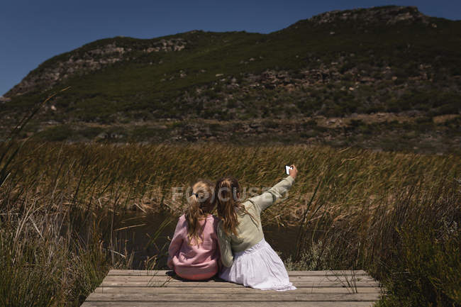 Vue arrière de frères et sœurs prenant selfie avec téléphone portable sur jetée — Photo de stock
