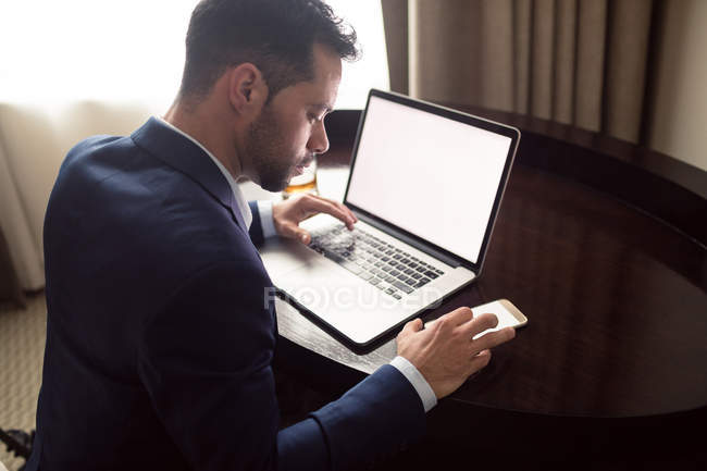 Empresario que utiliza el teléfono móvil mientras trabaja en el ordenador portátil en el hotel - foto de stock