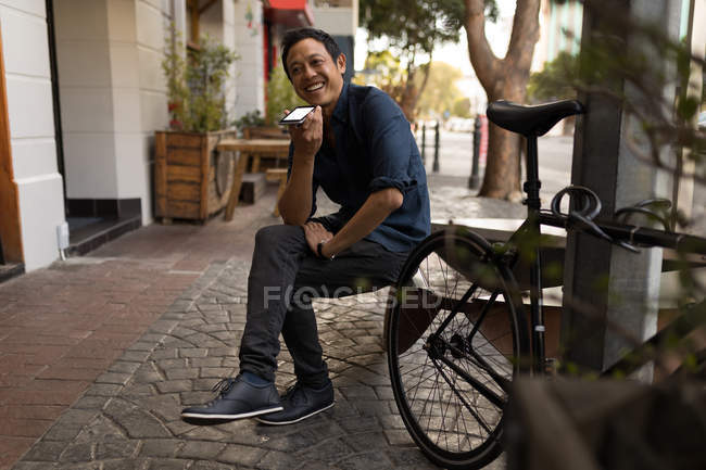 Homem sorridente em camisa azul falando no telefone celular no banco de café pavimento — Fotografia de Stock