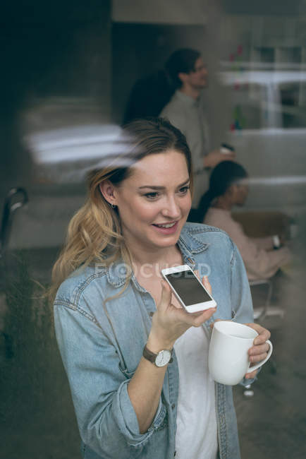 Female executive talking on mobile phone while having coffee in office — Stock Photo