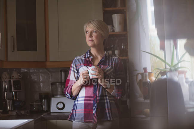 Задумчивая зрелая женщина пьет кофе на кухне дома — стоковое фото
