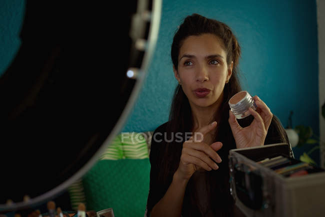 Beautiful woman holding, pointing at cosmetic accessory and looking away in front of mirror at home — Stock Photo
