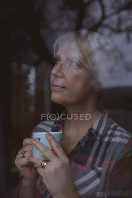 Mulher madura pensativo segurando xícara de café enquanto olha através da janela em casa — Fotografia de Stock