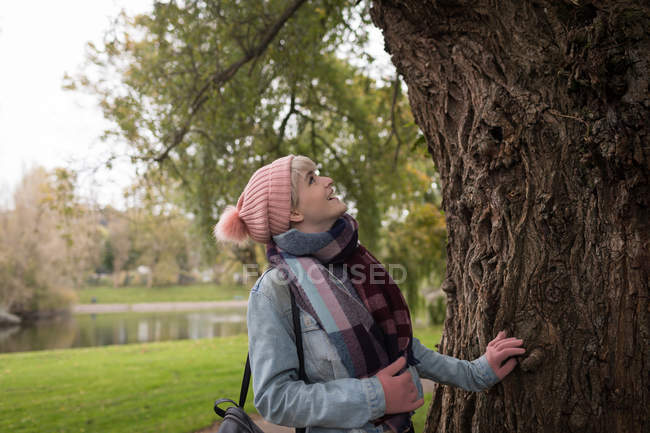 Mulher sorridente em roupas de inverno olhando para a árvore no parque — Fotografia de Stock