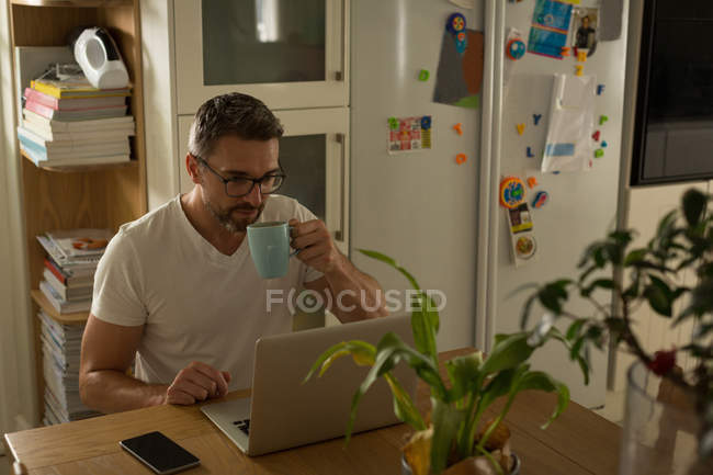 Мужчина пьет кофе дома, пользуясь ноутбуком — стоковое фото