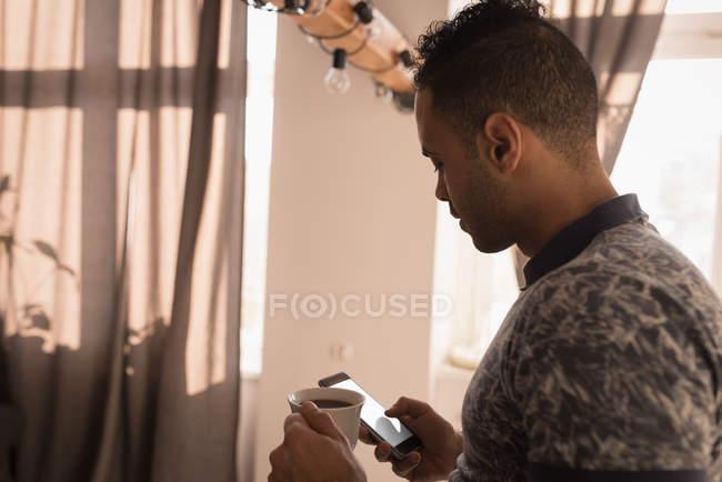 Homem usando telefone celular enquanto toma café na cozinha em casa . — Fotografia de Stock