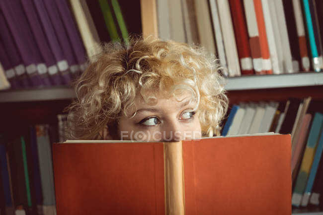 Jovem mulher escondendo seu rosto atrás do livro na biblioteca — Fotografia de Stock