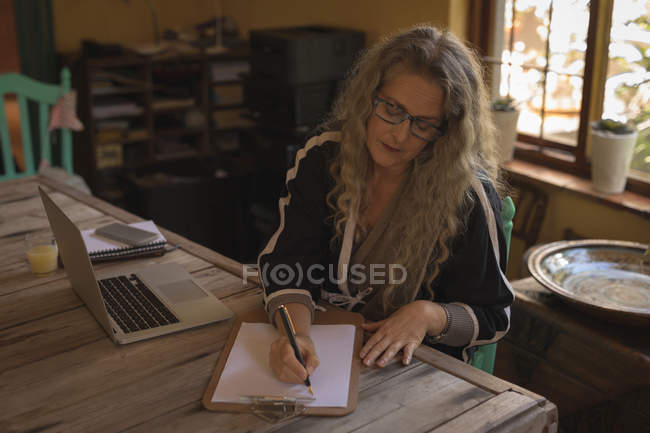 Mulher madura sentada na cadeira e escrevendo em papel em casa — Fotografia de Stock