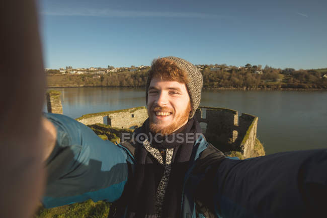 Счастливый турист-мужчина, стоящий у реки в сельской местности и позирующий для селфи — стоковое фото