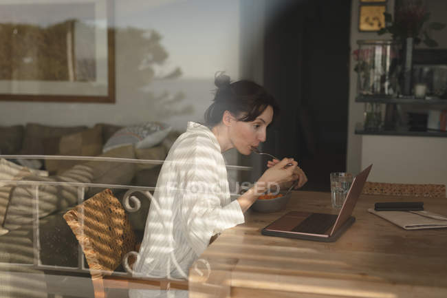 Jovem mulher tomando café da manhã enquanto olha para o laptop na sala de estar em casa — Fotografia de Stock