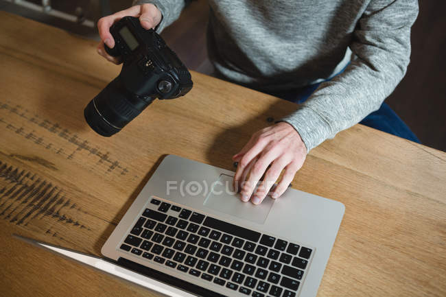Hombre usando el ordenador portátil mientras sostiene la cámara digital en casa - foto de stock