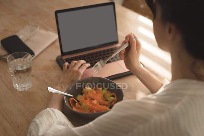 Молодая женщина ест завтрак, глядя в ноутбук в гостиной на дому — стоковое фото