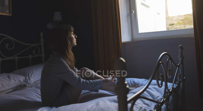 Женщина смотрит в окно, сидя дома на кровати — стоковое фото