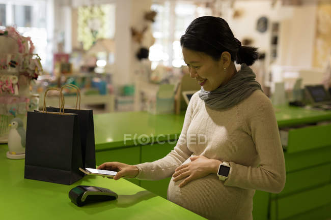 Mujer embarazada haciendo el pago a través del teléfono móvil en la tienda - foto de stock